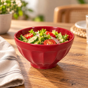 A red ceramic salad bowl filled with a fresh green salad, set on a wooden dining table with a striped napkin and blurred background elements.