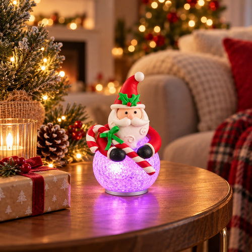 A festive Christmas ornament shaped like Santa Claus sitting on a glowing purple glittery orb, placed on a wooden table in a cozy, warmly lit living room with a Christmas tree and holiday decorations in the background.