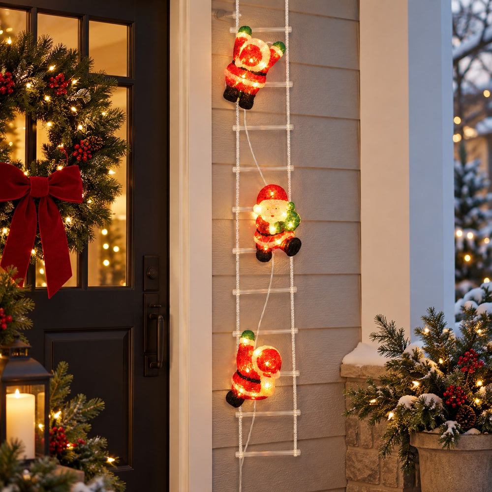 Three illuminated Christmas Santa Claus figures climbing a white ladder decoration on a house exterior at night, surrounded by holiday garlands, a wreath, lanterns, and a lit Christmas tree.