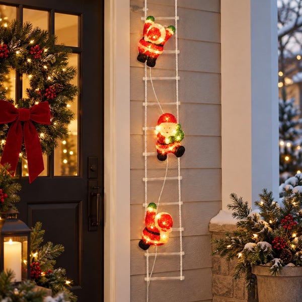 Three illuminated Santa Claus light figures climbing a white ladder decoration on a house exterior door frame, with holiday wreath, lantern, and snowy planters in the background.