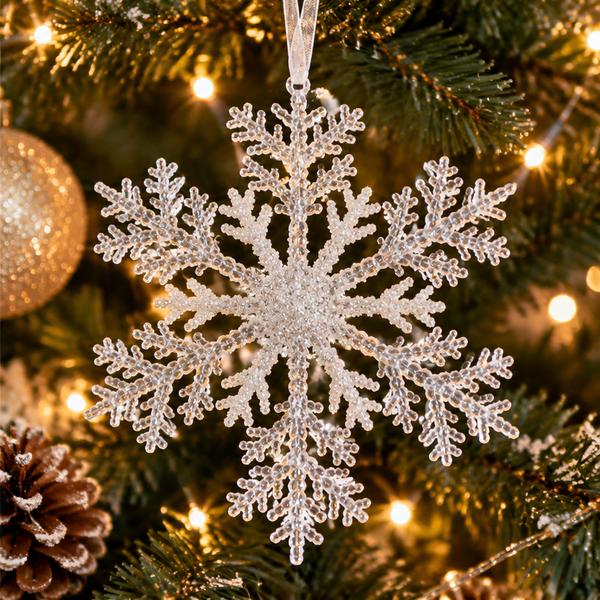 A large, intricate silver snowflake Christmas ornament hanging on a decorated Christmas tree with warm lights, pine needles, and a gold ornament in the background.