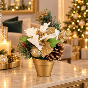 A festive holiday floral arrangement in a gold pot, featuring white poinsettias, pinecones, green pine needles, and glittery gold accents, displayed on a marble table in a warmly lit living room with a Christmas tree in the background.