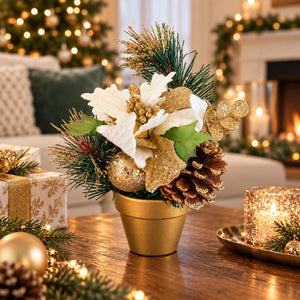 A festive holiday centerpiece in a small gold pot, featuring white poinsettia-style flowers, pinecones, green pine branches, and gold glitter accents, displayed on a wooden table in a warmly lit living room.