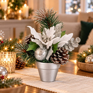 A festive Christmas centerpiece in a small silver pot, featuring silver and white poinsettias, pinecones, green pine branches, and glittery silver ornaments, styled on a table runner in a warmly lit living room.