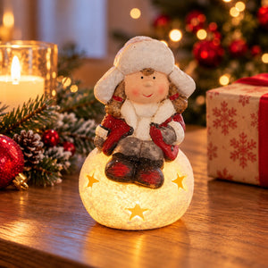 A Christmas snowman figurine glowing from within, sitting on a wooden table with a warm holiday background featuring a lit candle, pine branches, red ornaments, and a wrapped gift box.