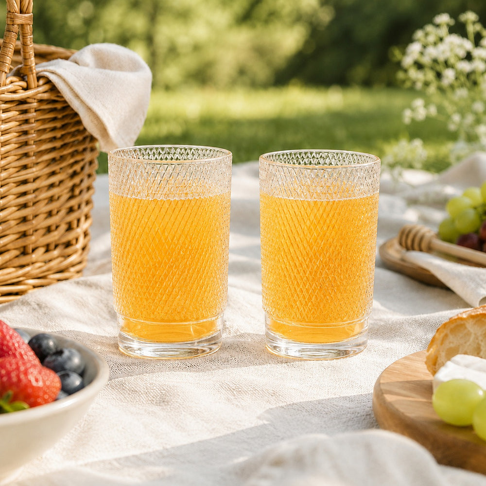 Two clear textured drinking glasses filled with orange juice on a white picnic blanket outdoors, with a wicker basket, fruit, and greenery in the background.