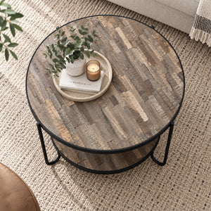 Round two-tier coffee table with a rustic wood top and black metal legs, styled in a living room setting with books, a potted plant, and a candle on the tabletop.
