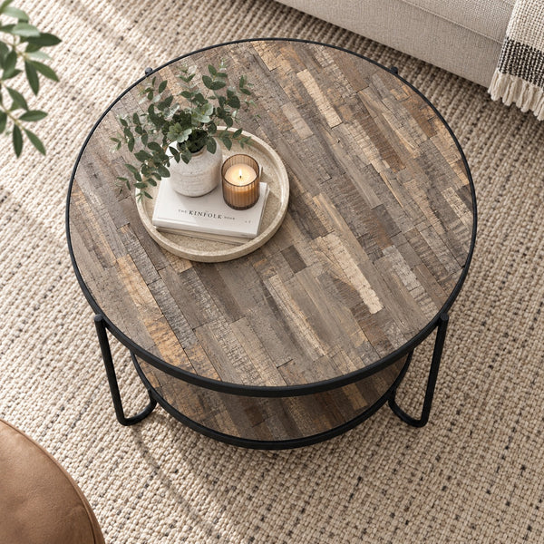 Round two-tier coffee table with a rustic wood top and black metal legs, styled in a living room setting with books, a potted plant, and a candle on the tabletop.