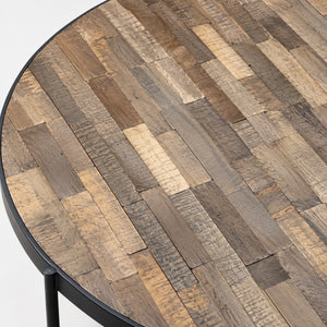 A close-up of a round table made from rustic reclaimed wood planks in mixed natural tones, with a black metal frame and leg visible at the lower left.