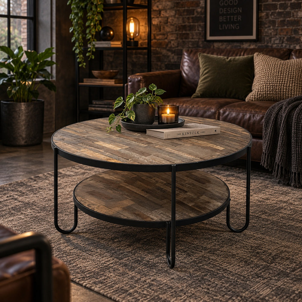 Round rustic coffee table with a two-tier wooden shelf and black metal legs, styled in a cozy living room next to a brown leather sofa.