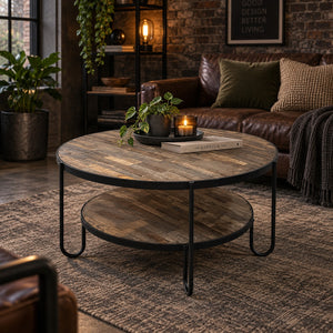 Round rustic coffee table with a two-tier wooden shelf and black metal legs, styled in a cozy living room next to a brown leather sofa.