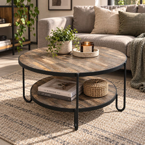 Round two-tier coffee table with a rustic wood top and black metal legs, styled in a living room setting with books, a potted plant, and a candle on the tabletop.
