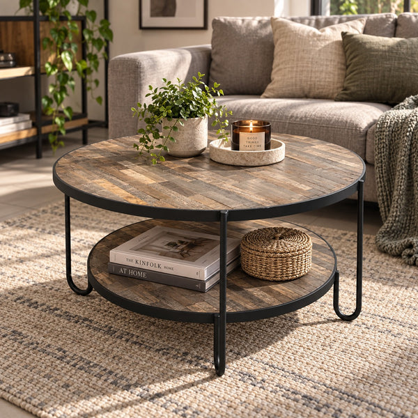 Round two-tier coffee table with a rustic wood top and black metal legs, styled in a living room setting with books, a potted plant, and a candle on the tabletop.