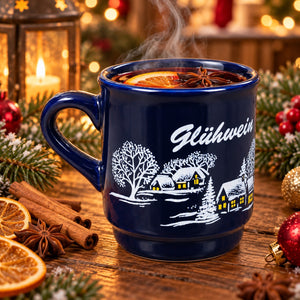 A dark blue ceramic mug with a white winter village illustration and the word 'Glühwein' on the side, filled with a steaming hot drink and garnished with orange slices and star anise, set on a wooden table surrounded by Christmas decorations.