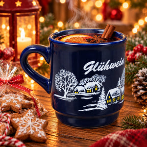 A dark blue ceramic mug with a white winter village illustration and the word 'Glühwein' on the side, filled with a hot holiday drink and garnished with orange slices, star anise, and cinnamon sticks. The mug sits on a wooden table surrounded by festive decorations including gingerbread cookies, pinecones, evergreen branches, and warm blurred lights in the background.