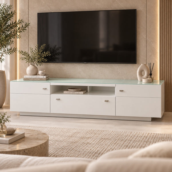 A modern white media console with a translucent green glass top, styled in a contemporary living room setting with a wall-mounted TV, decorative plants, and neutral-toned furniture.