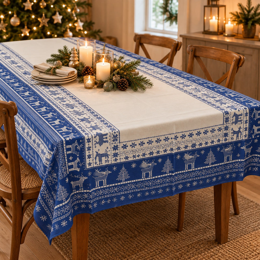 A blue and white Nordic-style Christmas table runner with reindeer and snowflake patterns, draped over a wooden dining table decorated for the holidays with candles, pinecones, and greenery.