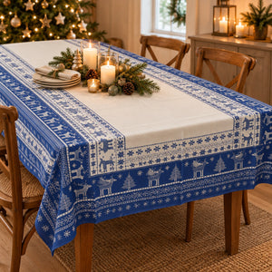 A blue and white Nordic-style Christmas table runner with reindeer and snowflake patterns, draped over a wooden dining table decorated for the holidays with candles, pinecones, and greenery.