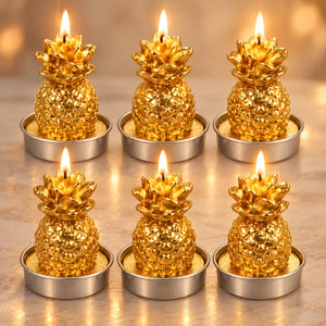 Six gold pineapple-shaped tea light candles with lit flames, arranged in two rows of three on a light marble surface with a warm bokeh background.