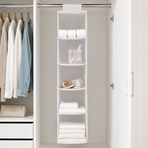 A white fabric closet organizer hanging from a metal rod inside a white closet, holding folded towels, rolled towels, a small jar, and a pair of sandals. Clothes are visible on hangers to the left.