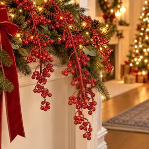 Red berry garland with green pine branches and warm white string lights draped over a white fireplace mantel, with a red ribbon bow on the left and a softly blurred decorated Christmas tree in the background.
