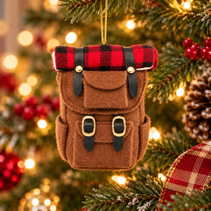 A brown felt backpack Christmas ornament hanging on a decorated Christmas tree with warm bokeh lights in the background.