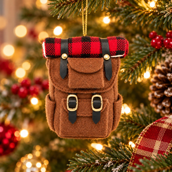 A brown felt backpack Christmas ornament hanging on a decorated Christmas tree with warm bokeh lights in the background.
