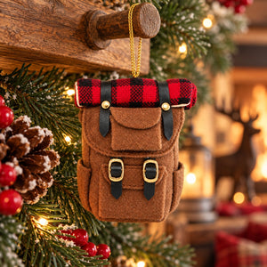 A brown felt Christmas ornament shaped like a small backpack or satchel, hanging from a wooden post. The top flap has a red and black buffalo plaid pattern, and the front has two black strap details with gold-tone buckle accents.