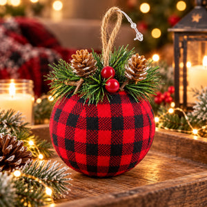 A round holiday ornament with a red and black buffalo plaid fabric cover, decorated with green pine branches, two pinecones, two red berries, and a jute hanging loop, set on a wooden tray with warm Christmas lights and candles in the background.