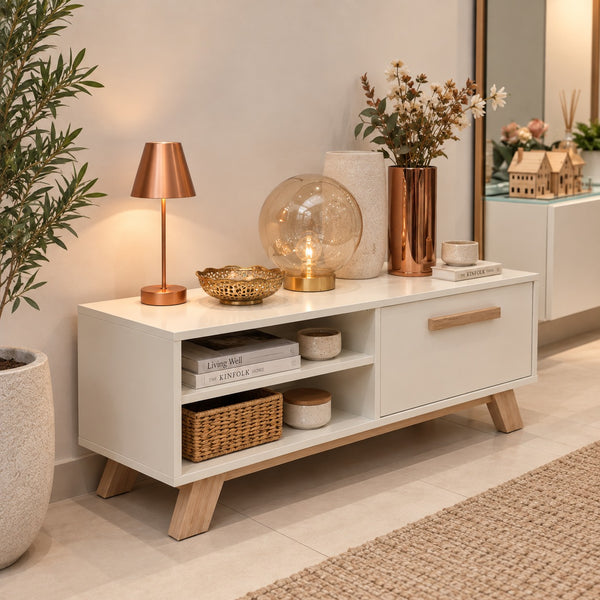 A modern white media console with light wood legs and handles, styled in a warm, contemporary room setting with decorative objects, a plant, and a mirror.