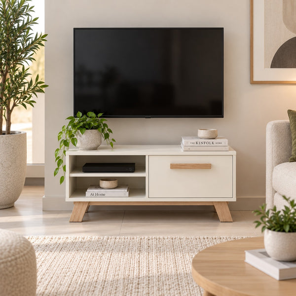 A modern white TV stand with light wood legs and a single wooden drawer handle, styled in a bright living room with a black television, potted plants, and home decor.