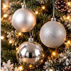 Three silver Christmas tree ornaments hanging on a decorated Christmas tree with warm lights. Two ornaments are matte and metallic silver, and one is reflective chrome silver.