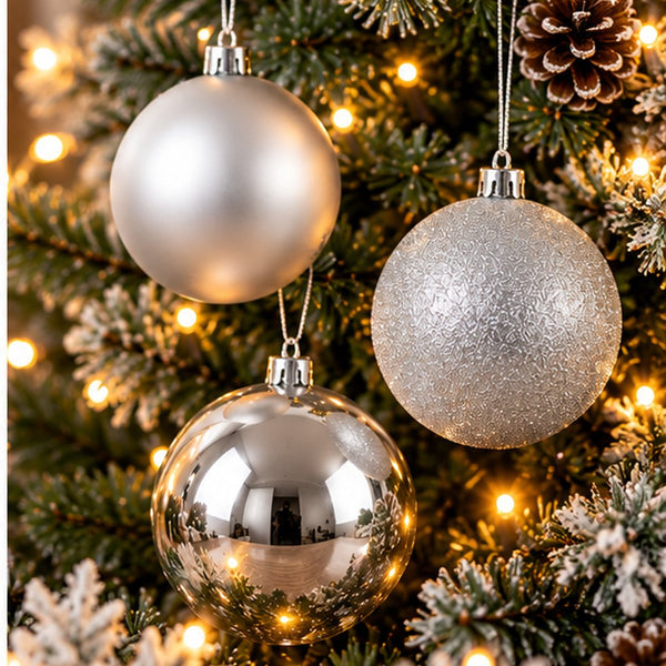 Three silver Christmas tree ornaments hanging on a decorated Christmas tree with warm lights. Two ornaments are matte and metallic silver, and one is reflective chrome silver.