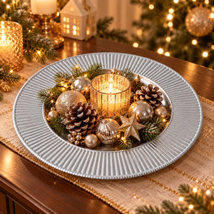 A round silver charger plate decorated with a festive Christmas centerpiece, including gold and silver ornaments, pinecones, green pine branches, and a lit candle, set on a wooden table with warm holiday lights in the background.