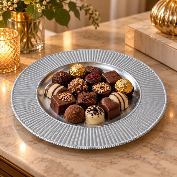 A silver ceramic plate with a fluted rim, filled with assorted chocolates and truffles, set on a beige marble countertop in a warm, decorated kitchen or dining area.