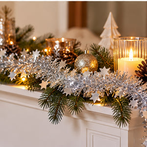 Silver tinsel garland with white star cutouts draped across a white fireplace mantel, styled with green pine branches, lit candles in glass holders, pinecones, and a gold ornament.