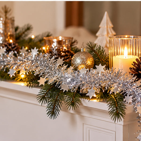 Silver tinsel garland with white star cutouts draped across a white fireplace mantel, styled with green pine branches, lit candles in glass holders, pinecones, and a gold ornament.