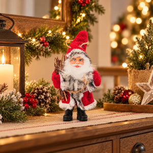 A festive Christmas Santa Claus figurine standing on a wooden dresser, surrounded by holiday decorations including a lantern, pine branches, red berries, and warm string lights in the background.
