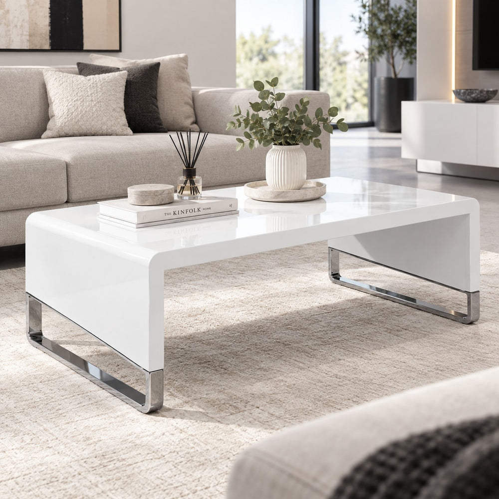 A modern white high-gloss rectangular coffee table with chrome metal legs, styled in a contemporary living room setting.