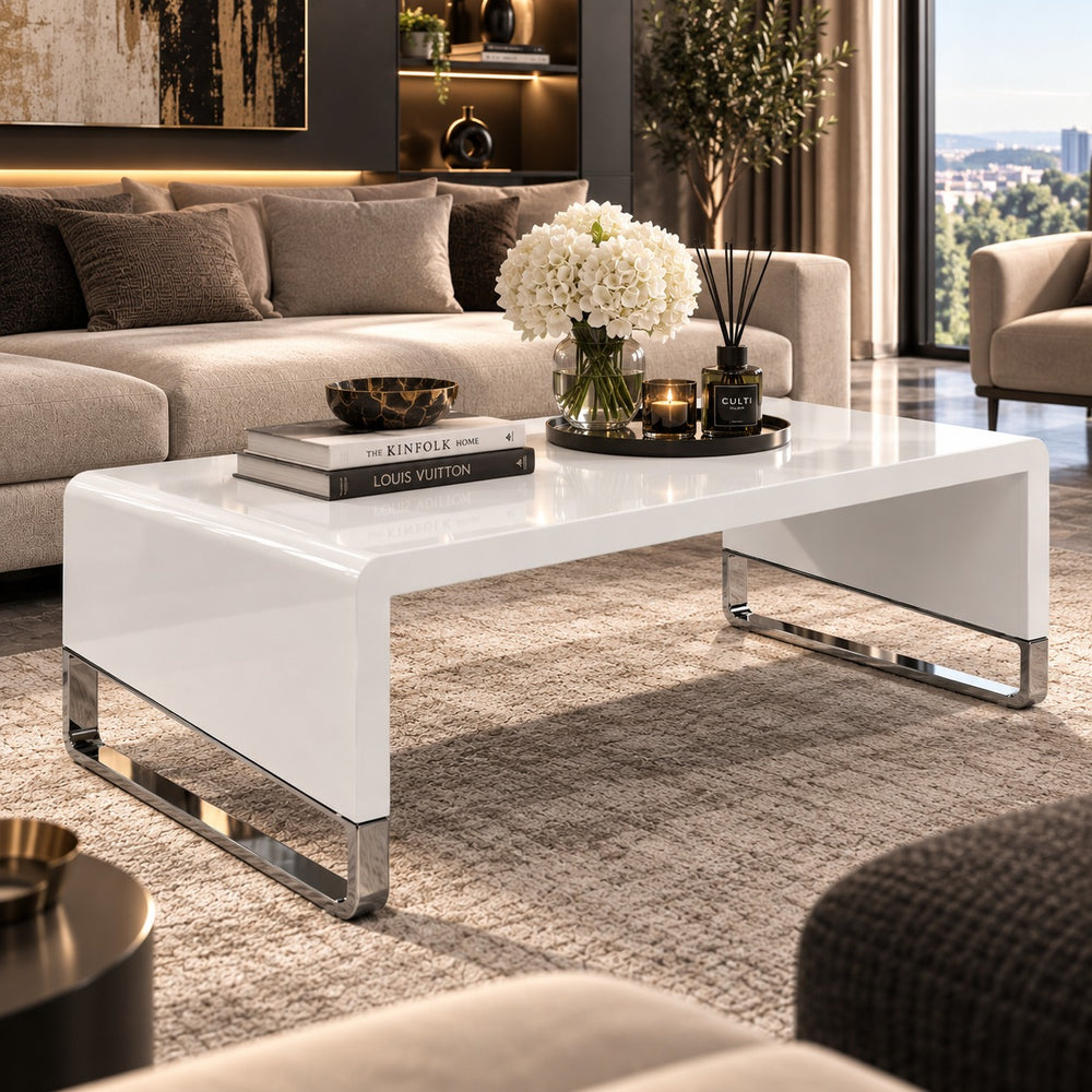 A modern white high-gloss rectangular coffee table with chrome metal legs, styled in a contemporary living room setting.