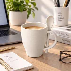 A beige ceramic coffee mug with a matching spoon, filled with coffee, set on a wooden desk surrounded by office items including a laptop, notebook, glasses, and a plant.