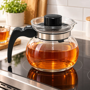 A clear glass teapot filled with amber tea sits on a black induction stovetop in a bright kitchen. The teapot has a black handle, black lid, and a silver metal collar around the glass.
