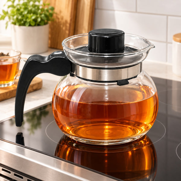 A clear glass teapot filled with amber tea sits on a black induction stovetop in a bright kitchen. The teapot has a black handle, black lid, and a silver metal collar around the glass.
