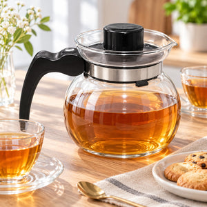 A clear glass teapot filled with amber tea sits on a wooden table, surrounded by matching glass cups, a spoon, cookies, and flowers in a bright, softly styled interior.