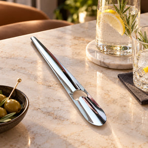 A polished chrome metal bar tool with a curved shaft and a circular cutout near one end, resting on a light beige marble table. The background features a glass of water with a lemon slice and rosemary, and a small bowl of olives.