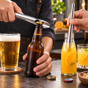 A close-up lifestyle shot of a person using a stainless steel bottle opener to open a brown glass beer bottle on a dark tabletop, with a glass of beer and a bottle of orange drink nearby.