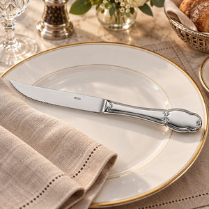 A silver metal steak knife with a decorative handle and 'MÉTRA' branding, resting on a white ceramic plate with a gold rim, set on a beige linen napkin in a styled dining setting.