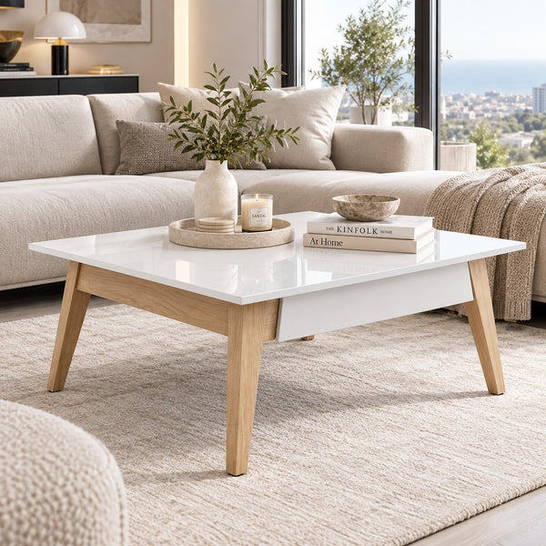 A modern rectangular coffee table with a glossy white top and light natural wood legs, styled in a bright living room with a sofa, rug, candles, books, and a vase.
