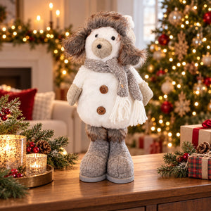 A plush dog doll dressed in a winter outfit with a white coat, gray scarf, brown fur trim, and gray boots, standing on a wooden table in a decorated Christmas living room.