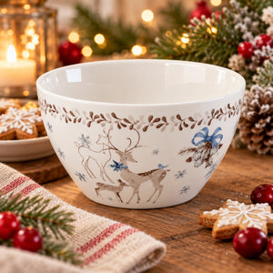 A white ceramic Christmas bowl decorated with brown deer, blue snowflakes, and a brown leaf border, set on a wooden table surrounded by holiday decorations like pine branches, red berries, gingerbread cookies, and a lit candle.